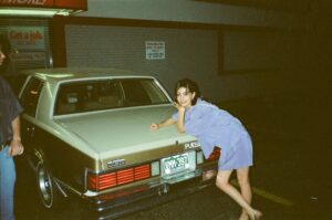 Woman in a blue dress leans on the trunk of a cream-colored vintage car at night in a parking lot in front of a storefront sign.