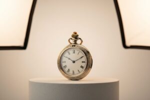 Ornate gold pocket watch on a white pedestal, lit by studio lights.