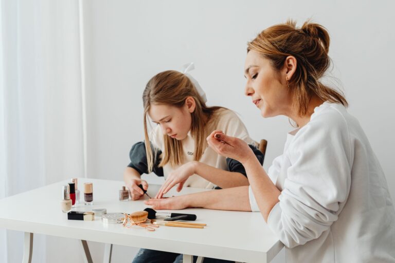 Woman and girl giving each other a manicure at a white table with assorted nail polishes and tools nearby.